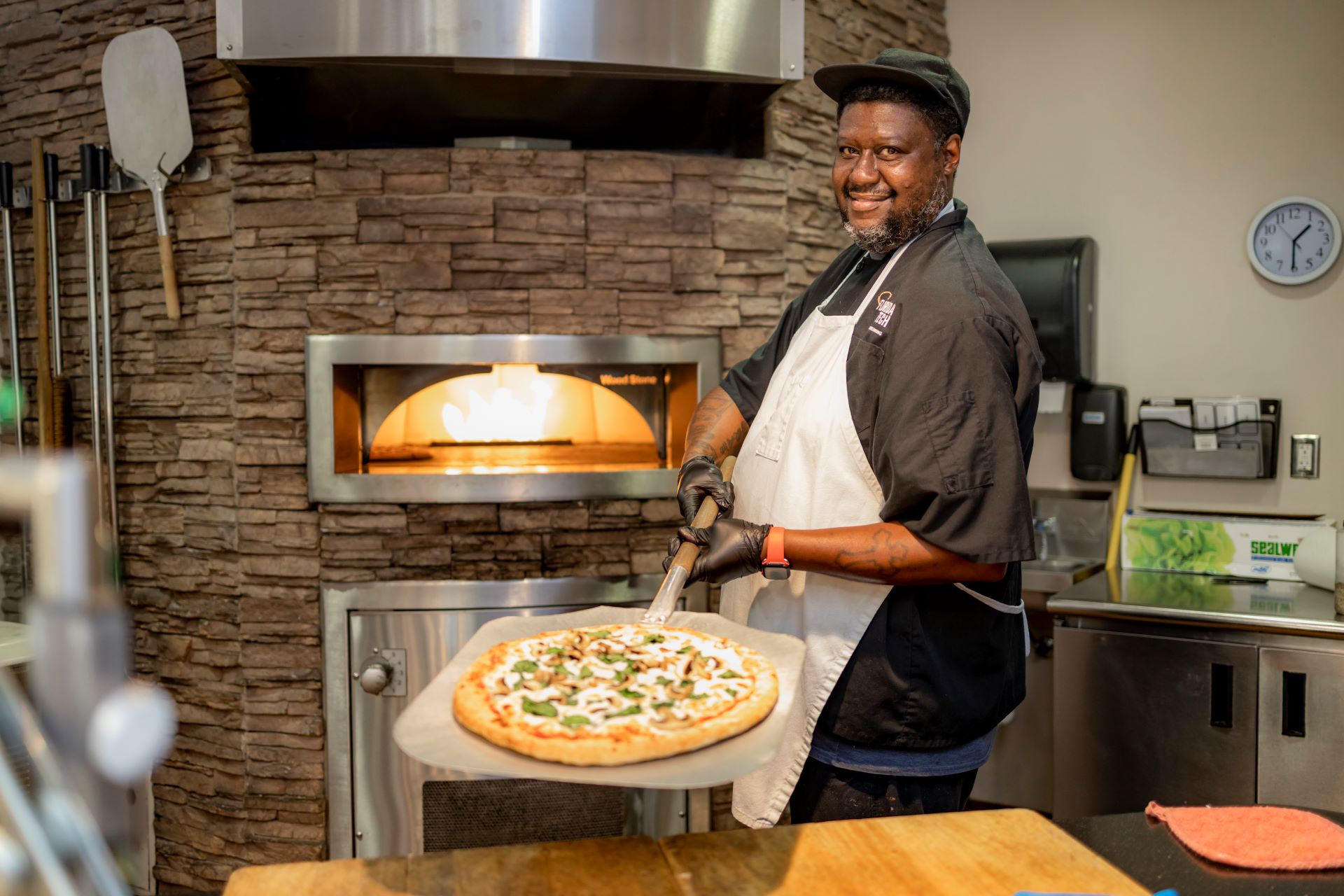 A chef smiling while removing a freshly baked pizza from a wood-fired oven in a restaurant kitchen